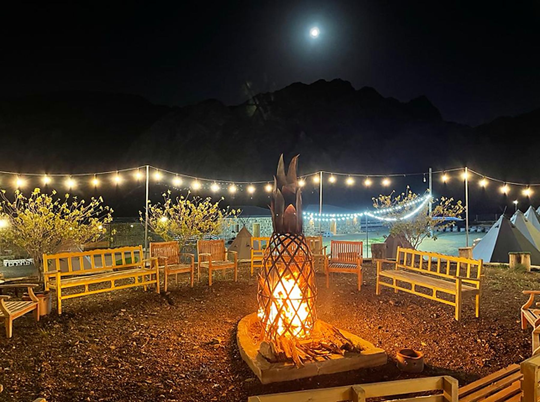 The full camp at night with a lit firepit, benches and strung lighting